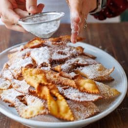 Dusting Angel Wing Cookies/ Polish Chrusciki with powdered sugar on a white platter