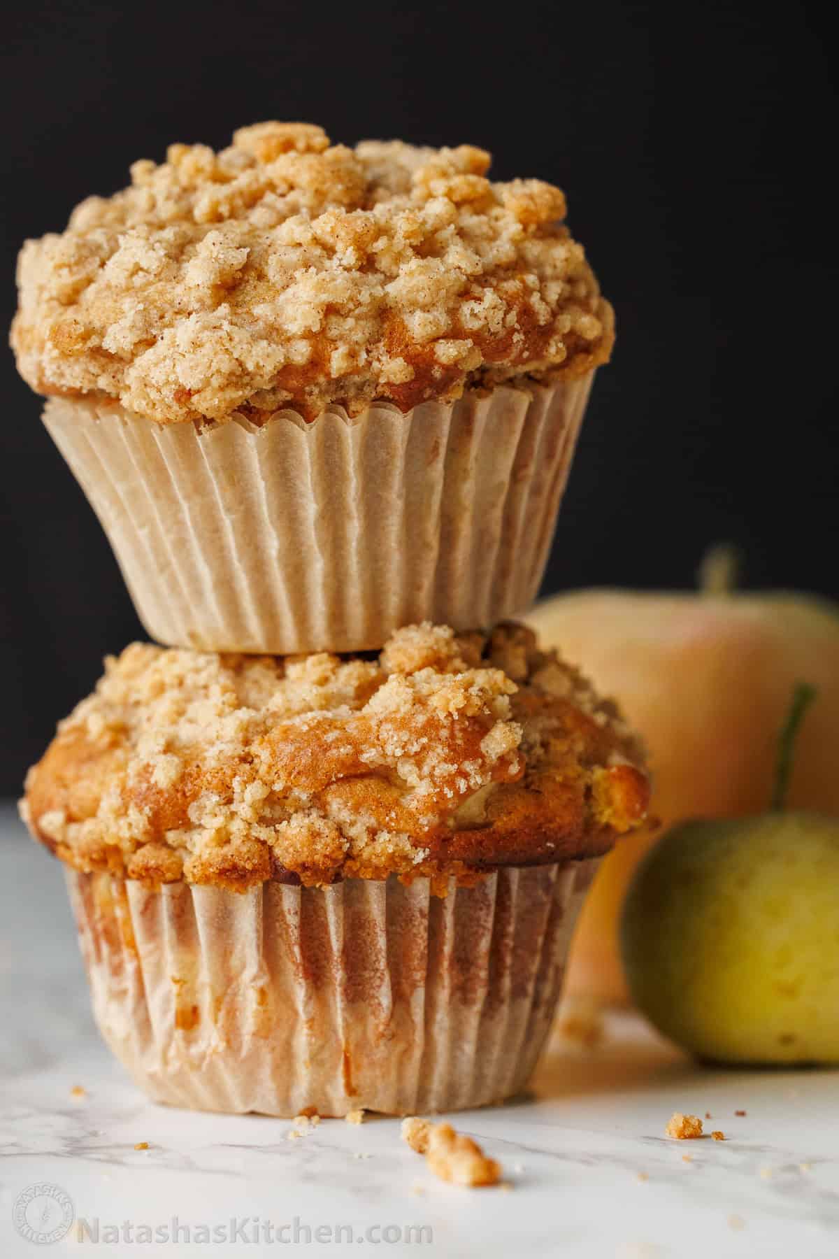 Two apple muffins stacked on a table with apples in the background