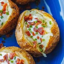 Baked potatoes on a blue plate, topped with sour cream, bacon, and chives.
