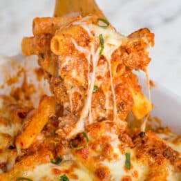 Baked ziti being scooped with a wooden spoon, showing the cheese pulls and layers of noodles, meat, cheese, and sauce.