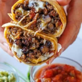 Beef burrito sliced in half to showcase the beef, melty Mexican cheese black beans, and Rotel tomatoes packed inside with bowls of salsa and guacamole toppings on the side.