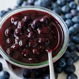 Homemade blueberry sauce in a glass jar with a spoon showcasing the whole blueberries. Fresh blueberries scattered on the counter around the jar.