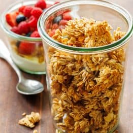 healthy Homemade granola in a glass jar near a yogurt parfait with raspberries and a spoon