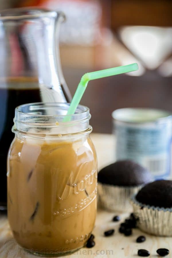 Iced coffee with condensed milk in a mason jar with a straw and cupcakes in the background 