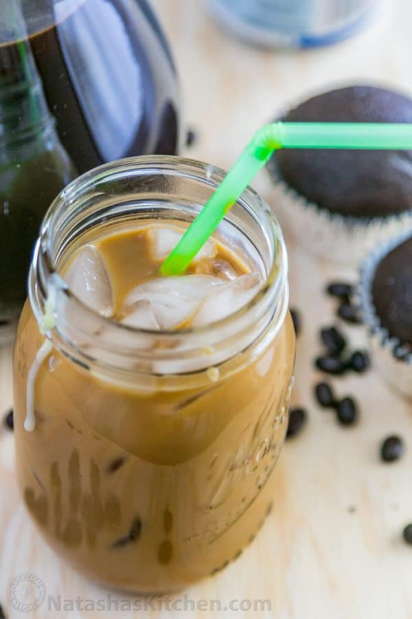 A close up of an iced coffee with condensed milk with cupcakes, coffee and coffee beans around the jar 