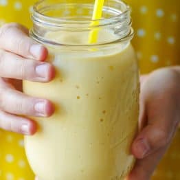 hands holding a yellow mango smoothie in a mason jar with yellow polka-dot background