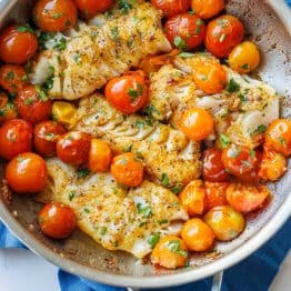 Flakey sauteed cod in a skillet with tomatoes and parsley garnish.