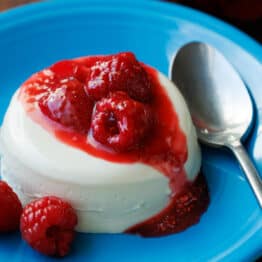 Panna Cotta with berry sauce drizzled on top, sitting on a blue plate with a spoon.
