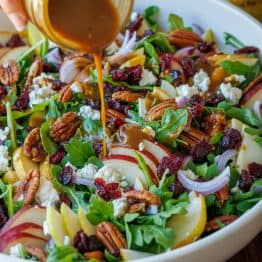 Pear Salad being dressed with Balsamic Vinaigrette in a white bowl, loaded with a variety of pears, red onion, goat cheese, cranberries, and toasted pecans