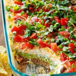 7-Layer Dip in a glass pan, showing each layer, refried beans, sour cream, cheese, tomatoes, green onion, guacamole, black olives with tortilla chips scattered around