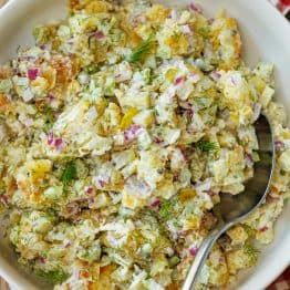 Smashed Potato Salad in a white bowl with a serving spoon, sitting on a red checkered towel.