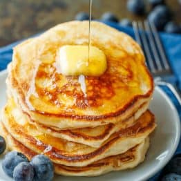 Sourdough Pancakes stacked on a plate with a pad of butter and maple syrup being poured over top.