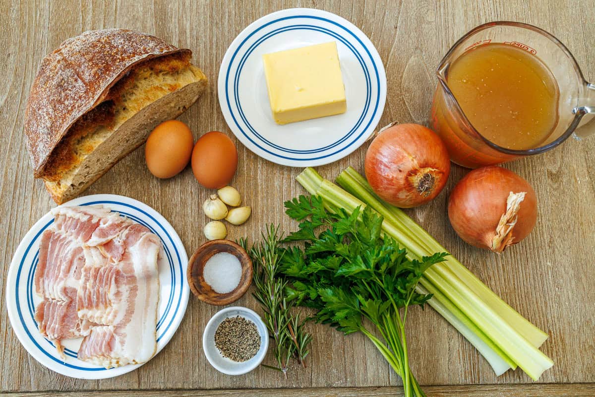 ingredients for the best Thanksgiving side dish with bread, eggs, garlic, butter, onions, celery, parsley, rosemary, salt and pepper, bacon, and chicken stock