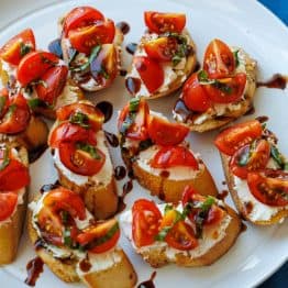 tomato goat cheese crostini on a white plate with cherry tomatoes and balsamic glaze on top